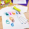 A white t-shirt with dinosaur stencils and airbrushed designs, along with an airbrush tool and markers, laid out on a wooden surface. Produktbild-Vorschau