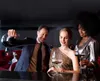 Three people are gathered around a bar, with one person pouring red wine from a decanter into a glass. The setting appears to be a sophisticated lounge or restaurant. Produktbild-Vorschau