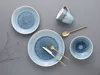 A top-down view of a blue stoneware dinnerware set arranged on a gray surface, with a sprig of dried foliage to the left. Náhľad obrázku produktu