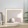 A lighted makeup mirror sits on a wooden dresser, reflecting a sofa and a plant. Podgląd zdjęcia produktu
