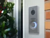 A smart doorbell mounted on a wall next to a window with plants. Produktbild-Vorschau