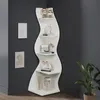 A white, wavy corner shelf unit is filled with decorative items and displayed in a room corner. Podgląd zdjęcia produktu