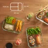 A top-down view of several glass meal prep containers filled with various sushi, shrimp, edamame, rice, avocado, and pickled ginger, arranged on a wooden surface. Anteprima dell’immagine prodotto