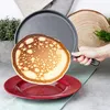 A hand is holding a cast iron frying pan with a pancake being flipped out of it onto a red plate. There is a plant and a wooden slice in the background. Produktbild-Vorschau