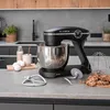 A black KLAMER kitchen mixer is shown on a kitchen counter with ingredients and accessories. Produktbild-Vorschau