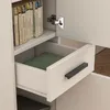 A close-up view of an open drawer in a white bookshelf, revealing a green book inside. A shelf above holds several books, and a cabinet hinge is visible on the right. Náhled obrázku produktu