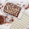 A chocolate cake with nuts in a glass container, next to a jar of chocolate spread and some hazelnuts and cinnamon sticks. Produktbild-Vorschau