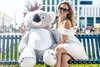 A woman sits on a bench next to a large teddy bear. Anteprima dell’immagine prodotto