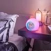 A digital alarm clock with a color-changing LED light sits on a nightstand next to a bed. Produktbild-Vorschau