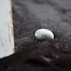 A close-up of a small, textured gray stone resting on a dark, furry surface, with a blurred white and gold object on the left. Produktbild-Vorschau