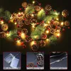 A Christmas garland with pine cones, pine needles, and red berries, illuminated by warm LED lights. The image also includes three smaller inset images showing a battery pack, the garland itself, and close-up of the lights. Produktbild-Vorschau