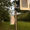 A weather station sensor unit mounted on a pole in an outdoor setting with trees and a grassy field in the background. Voir les images du produit