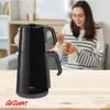 A woman is enjoying tea in a cozy living room setting, with a black Arzum tea maker prominently displayed on a wooden table in the foreground. Produktbild-Vorschau