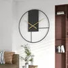 A modern wall clock is displayed on a white wall in a living room setting, next to a brown sofa and a bookshelf. Produktbild-Vorschau