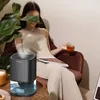 A woman sits in a chair, holding a drink and a book, with a dehumidifier on a table in the foreground. Produktbild-Vorschau
