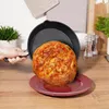 A hand is holding a cast iron pan with a round bread or pizza inside, tilting it over a red plate on a wooden table. Produktbild-Vorschau