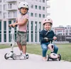Two children wearing helmets are on scooters in an outdoor setting with a modern building in the background. Náhled obrázku produktu
