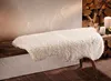 A fluffy beige faux sheepskin rug is draped over a rustic wooden bench in a cozy setting with a stack of firewood and candles in the background. Produktbild-Vorschau