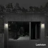 A modern black wooden house with a large window, illuminated by two wall lights at night. Produktbild-Vorschau