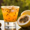 A close-up of a glass of passion fruit juice with ice and seeds, next to two passion fruits, with a blurred green background. Podgląd zdjęcia produktu