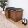 A wooden storage bench with a black cushion is shown in a room with a rug and a plant. Náhľad obrázku produktu