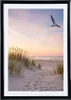 A framed picture of a beach scene with a seagull flying in the sky at sunset. Produktbild-Vorschau