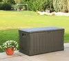 A brown plastic storage box sits on a patio next to a pot of white flowers, with a grassy yard and trees in the background. Produktbild-Vorschau