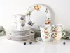 A set of porcelain cups, saucers, and plates with fruit illustrations, arranged on a white surface with some green fabric and blueberries. Náhľad obrázku produktu