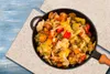 A close-up overhead view of a black skillet filled with a colorful vegetable stir-fry, placed on a textured beige cloth on a blue wooden surface. Náhled obrázku produktu