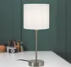 A white table lamp with a silver base and stem sits on a white desk in front of a dark green wall. In the background, out of focus, are some office supplies. Produktbild-Vorschau