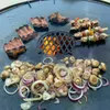 A large, round metal grill plate with food cooking on it, including ribs, skewers, mushrooms, and onions. Flames are visible underneath the grill. Produktbild-Vorschau