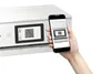 A hand holds a smartphone displaying a QR code, positioned next to a white printer with a QR code on its screen. Produktbild-Vorschau