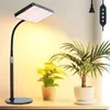 A grow light lamp is positioned on a desk next to two potted plants, with a power cord and controller visible in the background. Produktbild-Vorschau