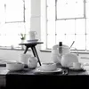 A table setting with white porcelain dishes, including bowls, cups, a teapot, and a sugar bowl, arranged in a bright room with large windows. Produktbild-Vorschau