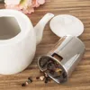 A white ceramic teapot with its lid and a metal tea infuser basket filled with dried tea leaves are arranged on a wooden surface. Some dried tea leaves are scattered on the surface around the infuser. Podgląd zdjęcia produktu