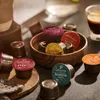 A collection of coffee capsules arranged in and around a wooden bowl, with a cup of espresso in the background. Anteprima dell’immagine prodotto