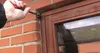 A person is using a screwdriver to attach a black mounting bracket to a brick wall next to a wooden window frame. Produktbild-Vorschau