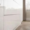 A close-up view of a white cabinet with two drawers, showing the glossy finish and a thin black line separating the drawers. The background includes a blurred curtain and a wooden floor. Produktbild-Vorschau