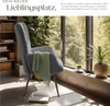 A comfortable armchair with a green throw blanket draped over it, placed near a large window with a view of trees. A book is open on the floor in front of the chair, and a cup of coffee sits on the windowsill. Produktbild-Vorschau