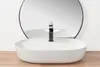 A white oval countertop sink with a black faucet is placed on a wooden surface in front of a large round mirror. Náhled obrázku produktu
