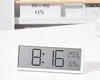 A digital clock displaying time, temperature, and humidity on a white desk. Produktbild-Vorschau