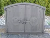 A cast iron oven door with decorative elements, set against a backdrop of green bushes and gravel. Náhľad obrázku produktu