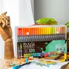 A set of dual-tip marker pens is displayed on a wooden table, with a wooden hand sculpture and some dried leaves. Produktbild-Vorschau
