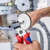 A person is using a pipe cutter to cut a gray pipe. The background shows some plumbing equipment. Produktbild-Vorschau