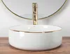 A white basin with a gold rim and a gold faucet is shown on a wooden surface, with a gold-rimmed mirror in the background. Produktbild-Vorschau