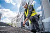A construction worker is drilling into a surface on a rooftop with a modern building in the background. Náhľad obrázku produktu
