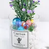 A close-up of a glass vase filled with white stones, colorful glass marbles, and artificial purple flowers. A label is attached to the vase. Náhľad obrázku produktu