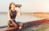 A woman in athletic wear drinks from a shaker bottle by a body of water. Produktbild-Vorschau