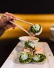 A hand using chopsticks to pick up a spring roll from a plate with other spring rolls and a dipping sauce. Produktbild-Vorschau
