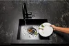 A person is rinsing a dirty plate with food scraps in a black kitchen sink under a black faucet. The countertop is black with white veining. Produktbild-Vorschau